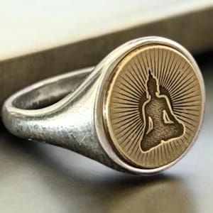 May include: A silver ring with a circular face featuring a seated Buddha figure. The Buddha is in a meditative pose, surrounded by sun rays. The ring has a brushed metal finish and a classic signet style.