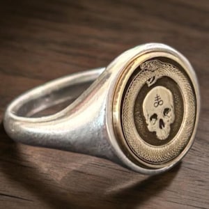 May include: A silver signet ring featuring a circular gold-toned inlay. The inlay displays an engraved ouroboros snake encircling a human skull with a sulfur symbol marked on the forehead. The ring has a polished metallic finish and a smooth band.