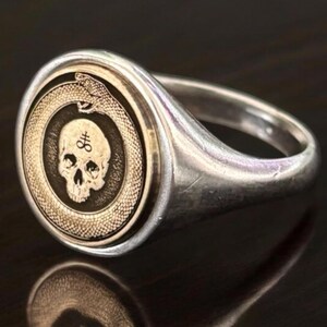 May include: A silver signet ring featuring a circular gold-toned inlay. The inlay displays an engraved ouroboros snake encircling a human skull. A small sulfur symbol is etched onto the forehead of the skull, centered within the ring face.