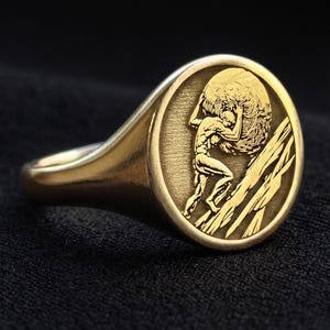 May include: A gold-toned signet ring featuring a circular face with an engraved depiction of a figure carrying a large sphere on their shoulders while climbing a rocky incline. The ring has a smooth, polished band and a detailed, recessed design.
