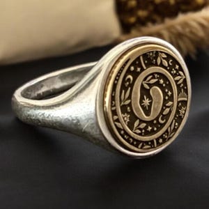 May include: A silver signet ring featuring a circular gold-toned inlay. The inlay displays an engraved spiral design surrounded by small botanical motifs, stars, and crescent moons. The ring band has a textured, matte finish with a rounded profile.