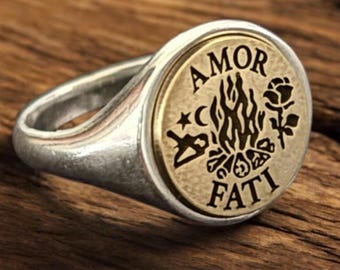Könnte beinhalten: Ein silberner Siegelring mit einer kreisförmigen Einlage in Messingoptik. Die Einlage ist mit dem Text AMOR FATI, einer zentralen Flamme, einem Halbmond, einem Stern und einer Rose graviert. Der Ring hat eine polierte Oberfläche und eine glatte Schiene.