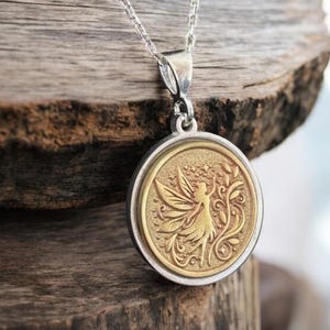 Puede incluir: Collar plateado con un colgante circular con un diseño de hada dorada. El hada tiene alas extendidas y está rodeada de detalles florales. El colgante cuelga de una delicada cadena plateada.