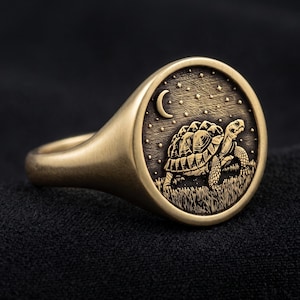 May include: A gold-toned signet ring featuring a circular face with an engraved design of a tortoise walking through grass under a night sky with a crescent moon and stars. The metal has a matte finish and a detailed, dark-etched relief pattern.