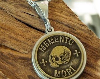 May include: A silver-toned necklace with a circular pendant. The pendant features a skull graphic and the words "MEMENTO MORI" in a circular design. The pendant is a bronze color with silver accents. The necklace is displayed on a wooden surface.