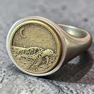Op de afbeelding: Een zilverkleurige zegelring met een ronde goudkleurige inleg. De inleg toont een getextureerde zeeschildpad die zwemt onder een maansikkel en sterren. De ring heeft een gladde, gepolijste band en een verhoogd, rond oppervlak met een metalen afwerking.