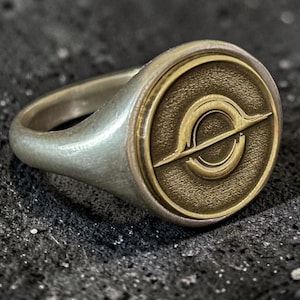 Op de afbeelding: Een zilverkleurige zegelring met een cirkelvormig vlak en een verzonken goudkleurige inleg. De inleg toont een reliëfafbeelding van een planeet met ringen of een hemellichaam met een horizontale lijn door het midden van de cirkel.