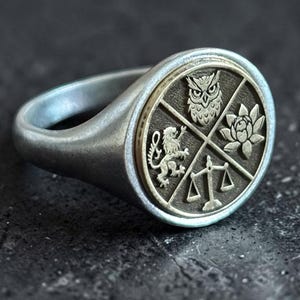 Peut inclure: Une bague chevalière en argent avec un plateau circulaire divisé en quatre quadrants par une ligne en forme de X. Les quadrants présentent des images en relief d'un hibou, d'une fleur de lotus, d'une balance et d'un lion sur fond métallique.