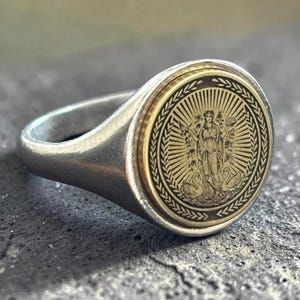 Può includere: Un anello a sigillo color argento con un inserto circolare color oro. L'inserto presenta una figura centrale incisa affiancata da due cigni, circondata da un motivo a raggiera e da un bordo decorativo a foglie di alloro.