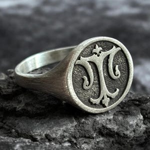 Pode incluir: Um anel de sinete em prata com uma face circular apresentando um monograma em relevo da letra T ornamentada. O metal possui um acabamento fosco escovado com áreas rebaixadas escurecidas ao redor do caractere central em destaque.