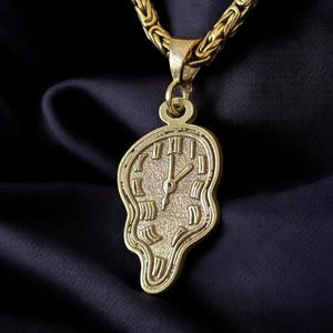 May include: Gold-toned necklace featuring a pendant shaped like a melting clock, a design inspired by Salvador Dalí's surrealist art. The chain is a thick, textured link style. The background is a dark, draped fabric.