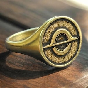 Puede incluir: Un anillo de sello dorado con una cara circular que presenta un fondo texturizado y un diseño central en relieve inspirado en un agujero negro. El anillo tiene una banda lisa y pulida con un motivo grabado que recuerda a un disco de acreción.