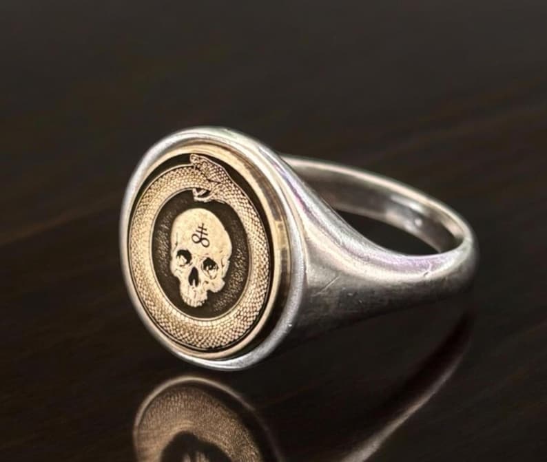 May include: A silver signet ring featuring a circular gold-toned inlay. The inlay displays an engraved ouroboros snake encircling a human skull. A small sulfur symbol is etched onto the forehead of the skull, centered within the ring face.