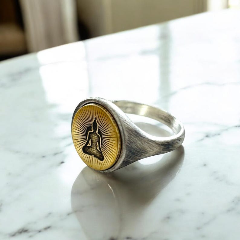 May include: A silver ring with a gold-colored face featuring a black silhouette of a seated Buddha. The ring has a brushed metal finish and a round face with sunburst rays emanating from the Buddha.
