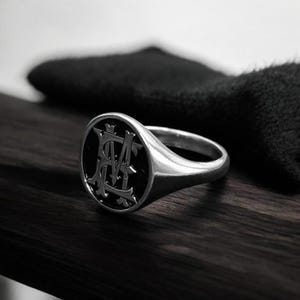 K&ouml;nnte beinhalten: Ein silberner Siegelring mit schwarzem, ovalem Gesicht. Das Gesicht zeigt ein verziertes, verschlungenes Monogramm in hellerer Silberfarbe. Der Ring ist vor einer dunklen Holzoberfl&auml;che und einem schwarzen, strukturierten Hintergrund platziert.