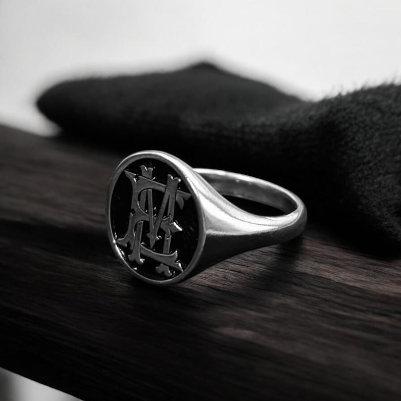 K&ouml;nnte beinhalten: Ein silberner Siegelring mit schwarzem, ovalem Gesicht. Das Gesicht zeigt ein verziertes, verschlungenes Monogramm in hellerer Silberfarbe. Der Ring ist vor einer dunklen Holzoberfl&auml;che und einem schwarzen, strukturierten Hintergrund platziert.