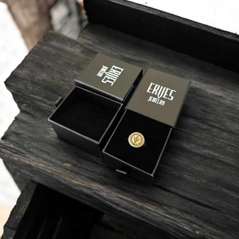 K&ouml;nnte beinhalten: Zwei offene schwarze Schmuckschatullen mit dem Text "ERVES JEWELRY" in Wei&szlig;. Eine Schatulle enth&auml;lt einen goldenen Ring mit einem kreisf&ouml;rmigen Emblem. Die Schatullen liegen auf einer dunklen, strukturierten Holzoberfl&auml;che.