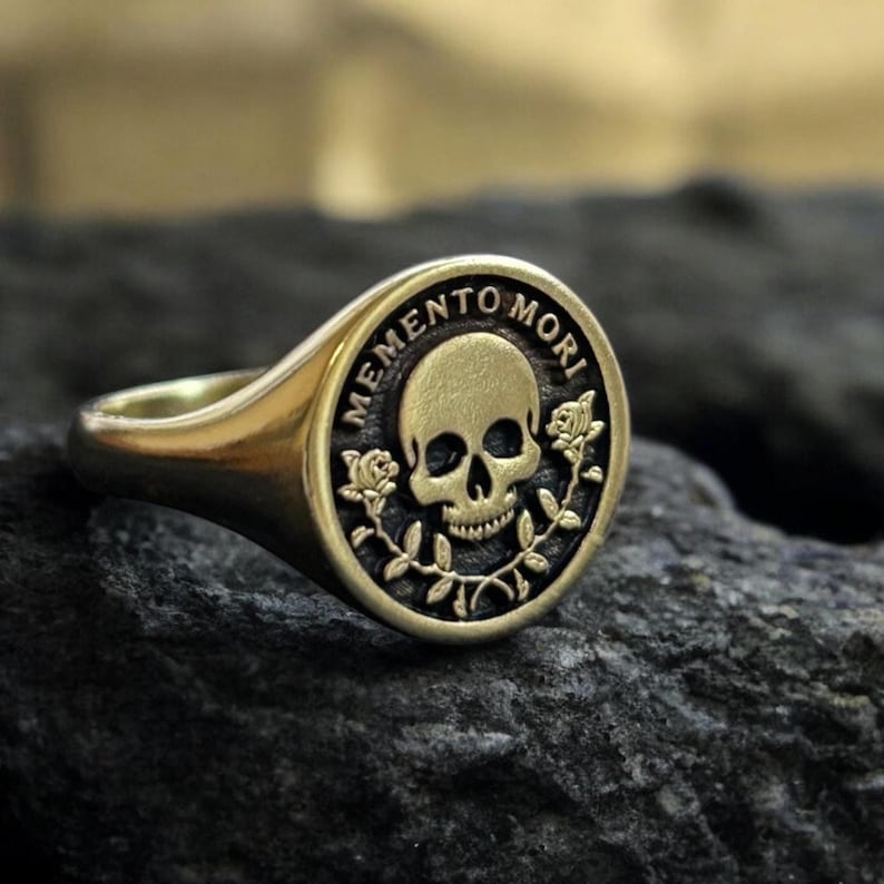 May include: A gold signet ring with a skull and roses design. The ring features the inscription "Memento Mori" around the skull.