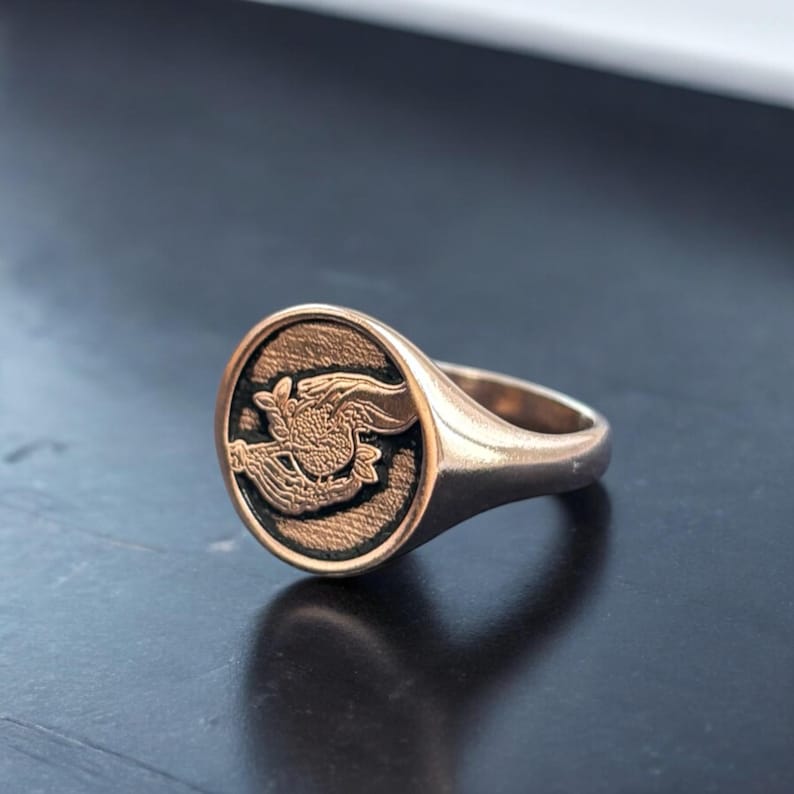 Op de afbeelding: Een gouden zegelring met een gedetailleerd gegraveerd ontwerp van een mythisch wezen met vleugels en een staart.