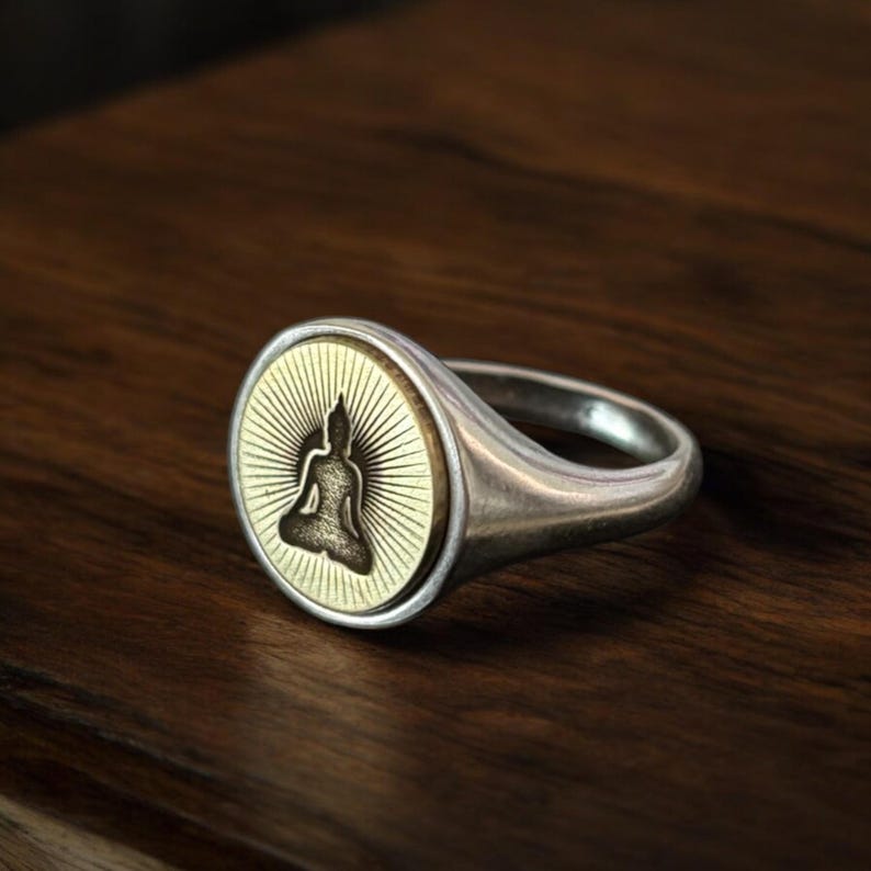 May include: A silver ring with a gold-colored circular face featuring a seated Buddha figure. The Buddha is centered within a sunburst design. The ring has a polished finish and a classic signet style.