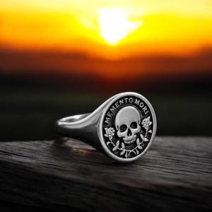 May include: A silver signet ring with a black skull and crossbones design. The ring has the words "Memento Mori" engraved around the skull.