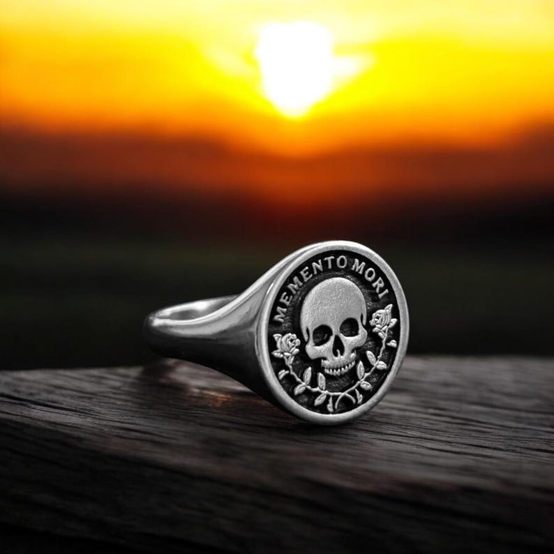May include: A silver signet ring with a black skull and crossbones design. The ring has the words "Memento Mori" engraved around the skull.