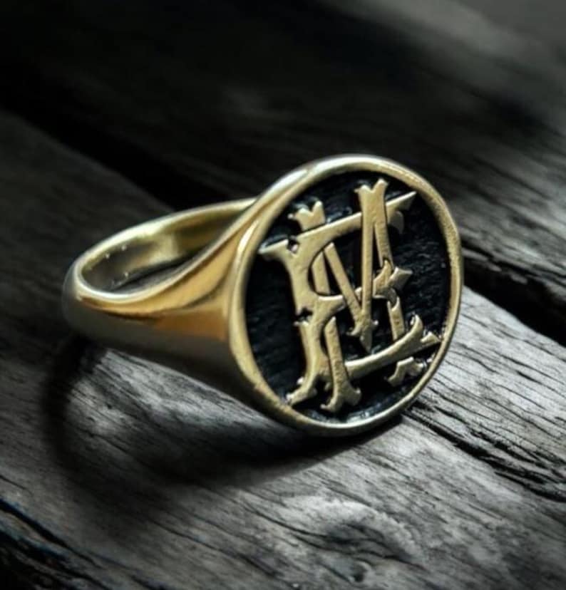 K&ouml;nnte beinhalten: Ein goldfarbener Siegelring mit schwarzem, ovalem Gesicht. Das Gesicht zeigt ein verziertes, verschlungenes Monogramm in Gold. Der Ring liegt auf einer dunklen, strukturierten Holzoberfl&auml;che.