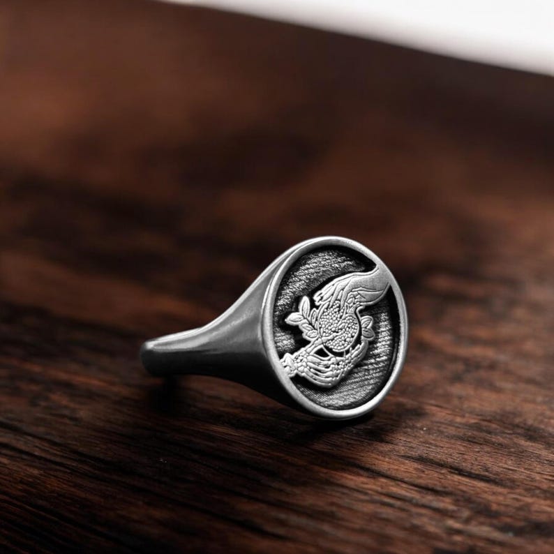 Op de afbeelding: Een zilveren zegelring met een gedetailleerd ontwerp van twee handen die een bol vasthouden. De ring ligt op een houten achtergrond.