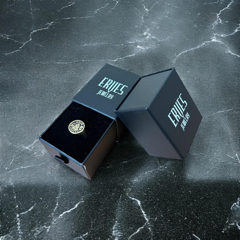 May include: A black ring box with the words "Erves Jewelry" printed on the top and side. The box is open and a gold ring with a design is inside.