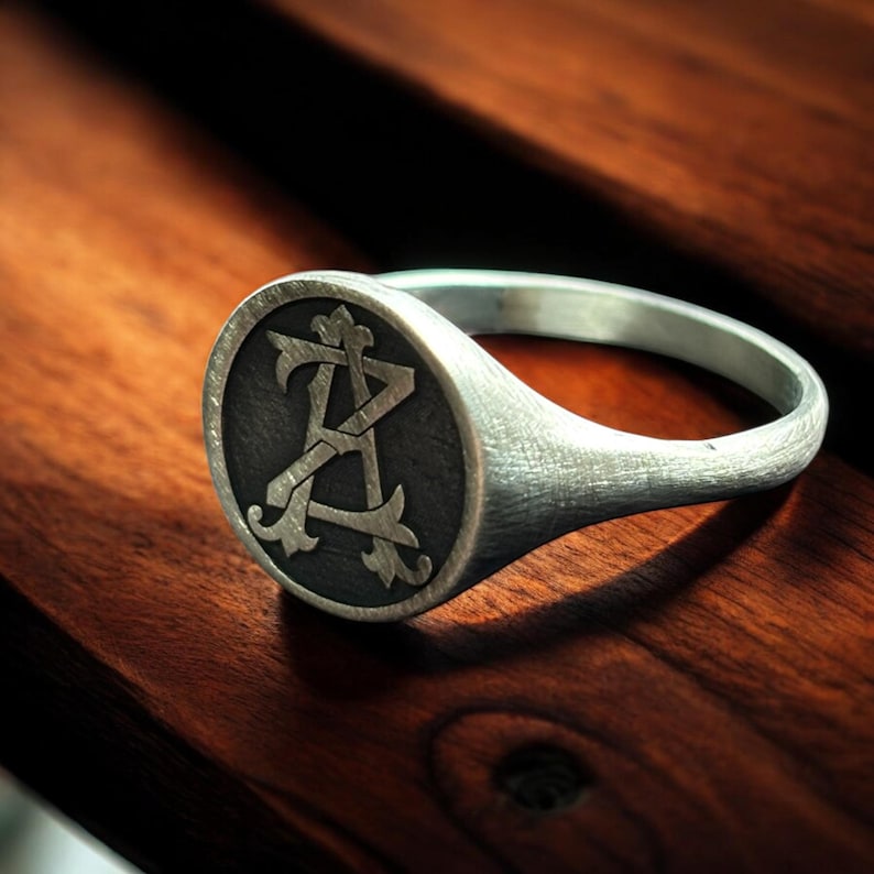 K&ouml;nnte beinhalten: Ein silberner Siegelring mit einer dunkelgrauen, kreisf&ouml;rmigen Oberfl&auml;che. Die Oberfl&auml;che zeigt ein verziertes Design der Buchstaben "A" und "Z", die miteinander verschlungen sind. Der Ring liegt auf einer dunklen Holzoberfl&auml;che, die den metallischen Glanz des Rings hervorhebt.