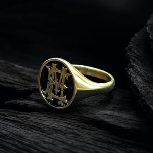 K&ouml;nnte beinhalten: Ein goldener Siegelring mit schwarzem ovalem Gesicht. Das Gesicht zeigt ein verziertes, verschlungenes Monogramm in hellerer Goldfarbe. Der Ring ist vor einem dunklen, strukturierten Hintergrund platziert.