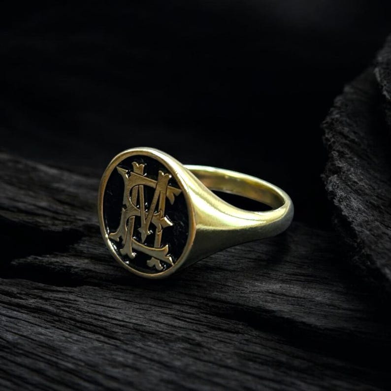 K&ouml;nnte beinhalten: Ein goldener Siegelring mit schwarzem ovalem Gesicht. Das Gesicht zeigt ein verziertes, verschlungenes Monogramm in hellerer Goldfarbe. Der Ring ist vor einem dunklen, strukturierten Hintergrund platziert.