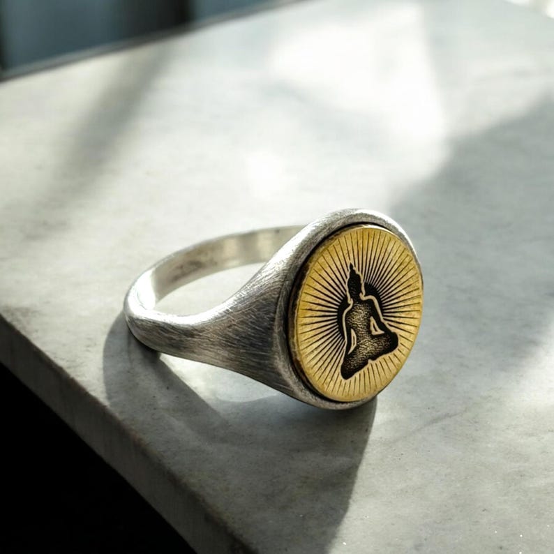 May include: A silver ring with a gold-colored, circular face featuring a black silhouette of a seated Buddha. The face has a sunburst design radiating from the Buddha. The ring has a brushed metal finish.