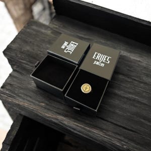 May include: Two open black jewelry boxes with the text "ERVES JEWELRY" in white. One box contains a gold ring with a circular emblem. The boxes sit on a dark, textured wooden surface.