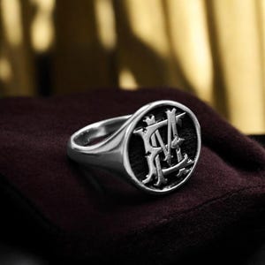 K&ouml;nnte beinhalten: Ein silberner Siegelring mit dunkler, strukturierter Oberfl&auml;che. Die Oberfl&auml;che zeigt ein verziertes Monogramm-Design in hellerer Silberfarbe. Der Ring ist vor einem dunkelroten Hintergrund platziert.