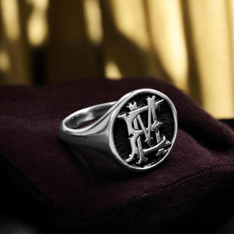 K&ouml;nnte beinhalten: Ein silberner Siegelring mit dunkler, strukturierter Oberfl&auml;che. Die Oberfl&auml;che zeigt ein verziertes Monogramm-Design in hellerer Silberfarbe. Der Ring ist vor einem dunkelroten Hintergrund platziert.