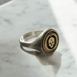 May include: A silver-toned signet ring featuring a circular gold-toned inlay. The inlay displays a detailed engraving of a human skull positioned beneath a sulfur symbol, all encircled by a textured serpent motif forming a continuous ring shape.