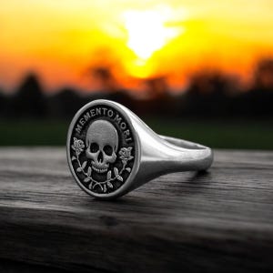 May include: A silver signet ring with a black skull and roses design. The ring features the inscription "Memento Mori" around the skull.