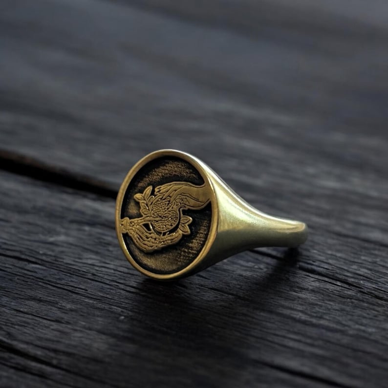 Op de afbeelding: Een gouden zegelring met een gedetailleerd gegraveerd ontwerp van een hand die een tak met bladeren vasthoudt. De ring is ovaalvormig en heeft een gladde band.