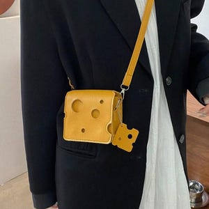 以下が含まれることがあります： スイスチーズの形をした小さな黄色のクロスボディバッグ。チーズのくさび形のチャーム付き。