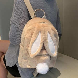 以下が含まれることがあります： ウサギの形をしたベージュのぬいぐるみバックパック。白い内側のある大きな直立した耳、小さなジッパーポケット、ふわふわの白い尻尾が特徴です。バッグには調節可能なストラップと上部のハンドルが付いています。