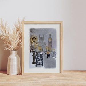 May include: A framed watercolor print depicts a snowy scene of London, featuring Big Ben and the Houses of Parliament. The artwork shows a street with benches and lampposts, with two figures walking along a snow-covered path. The frame is light-colored.