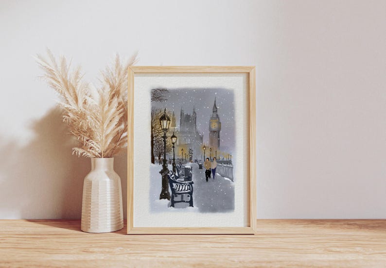 May include: A framed watercolor print depicts a snowy scene of London, featuring Big Ben and the Houses of Parliament. The artwork shows a street with benches and lampposts, with two figures walking along a snow-covered path. The frame is light-colored.