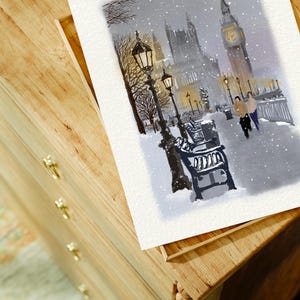 May include: Watercolor painting of a snowy London scene featuring Big Ben and the Houses of Parliament. The artwork depicts a winter landscape with falling snow, street lamps, and benches. The painting is on a white paper, resting on a wooden surface.