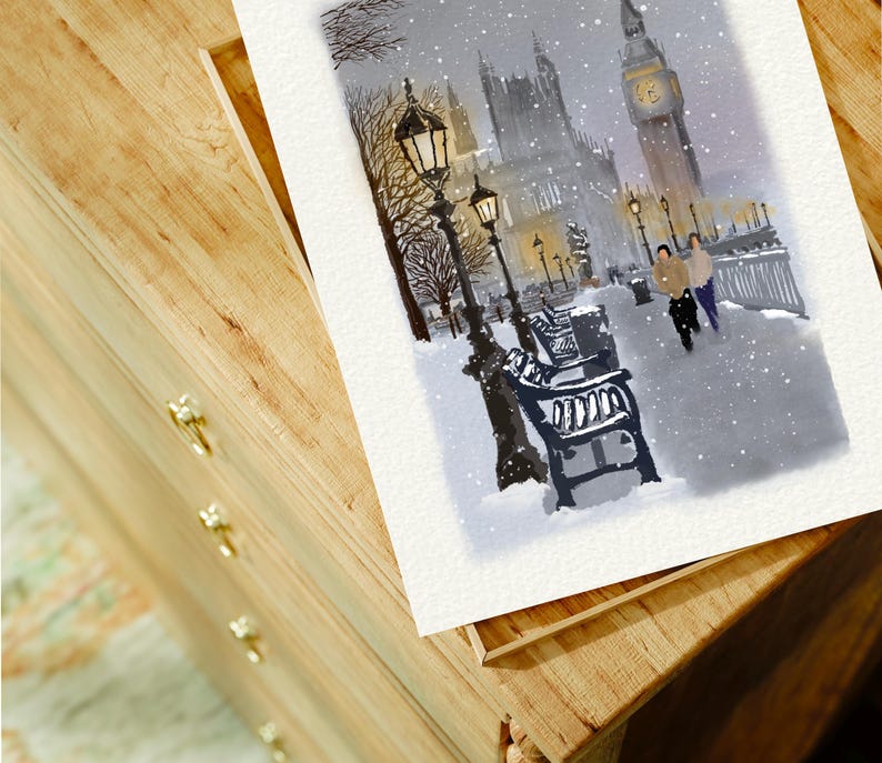 May include: Watercolor painting of a snowy London scene featuring Big Ben and the Houses of Parliament. The artwork depicts a winter landscape with falling snow, street lamps, and benches. The painting is on a white paper, resting on a wooden surface.