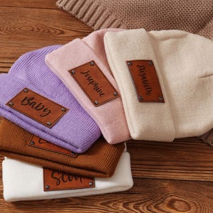 May include: A collection of folded beanies in various colors, including lavender, pink, brown, and white. Each beanie features a brown leather patch with text, such as "Baby", "Josephine", "Elizabeth", and "Scot". The beanies are arranged on a wooden surface.