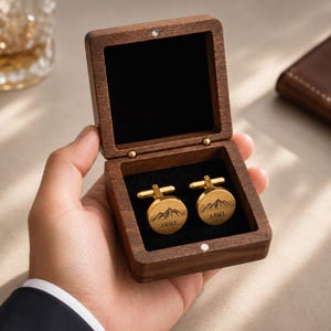 Peut inclure: Une paire de boutons de manchette ronds en métal doré avec un motif de chaîne de montagnes gravé et le nom ARIEL, présentés dans une boîte carrée en bois brun foncé avec une doublure intérieure en velours noir et un couvercle à charnière.