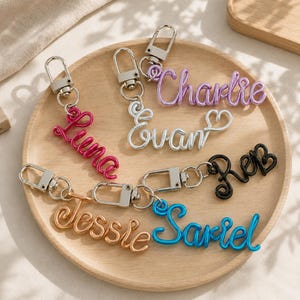 May include: Five custom wire name keychains with silver-toned lobster clasps. The names Luna in pink, Charlie in purple, Evan in silver with a heart, Reb in black, Jessie in copper, and Sariel in blue are formed from continuous wire script.