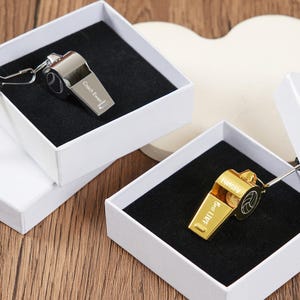 Op de afbeelding: Twee metalen fluitjes, één zilver en één goud, gepresenteerd in witte dozen met zwarte fluwelen inzetstukken. Op het zilveren fluitje staat de tekst "Coach Evan". Het gouden fluitje heeft de tekst "Best Life" en "LUMSEY".