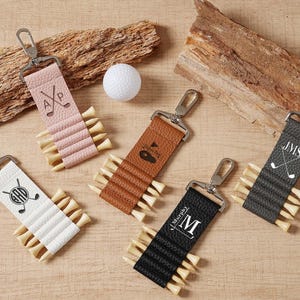 Op de afbeelding: Een verzameling gepersonaliseerde golf tee-houders in verschillende kleuren, waaronder roze, bruin, wit, zwart en grijs. Elke houder heeft een metalen sluiting en wordt gecombineerd met houten golf tees. Golfballen zijn ook aanwezig.